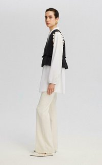 Chaleco negro cortado con dobladillo con volantes sobre una camisa blanca de manga larga, combinado con pantalones blancos de corte acampanado y zapatos planos con punta.
