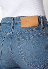 Jeans de mezclilla azul con un parche de cuero con la inscripción "FRAME" en relieve. La tela tiene una textura desgastada, con costuras visibles y bolsillos traseros.