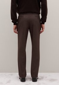 Pantaloni marroni su misura in tessuto strutturato, con taglio dritto, due tasche posteriori con chiusura a bottone, indossati con scarpe nere.