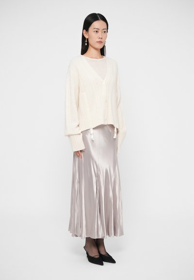 Krémový krátký svetr s copánkovým vzorem, hedvábně vypadající midi sukně světle šedé barvy, černé špičaté boty, modelka s dlouhými černými vlasy stojí před jednobarevným pozadím.