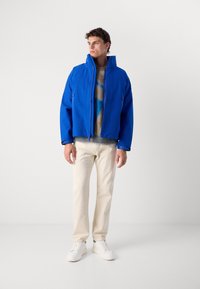 Giacca cropped blu con alto colletto, zip frontale e tasche laterali. Indossata sopra un maglione beige e pantaloni crema, completata da sneakers bianche.