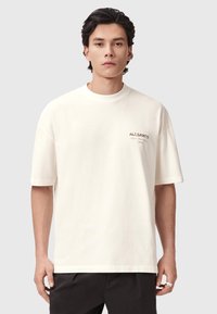 Μπλουζάκι από βαμβάκι σε κρεμ χρώμα με στρογγυλή λαιμόκοψη και κοντά μανίκια, με ένα διακριτικό λογότυπο τοποθετημένο στην αριστερή πλευρά.