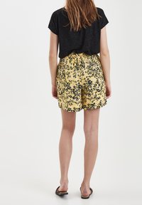 Keltaiset shortsit mustalla kukkakuviolla, joustavalla vyötäröllä ja rentolla istuvuudella. Yhdistetty mustaan lyhythihaiseen toppiin ja sandaaleihin.