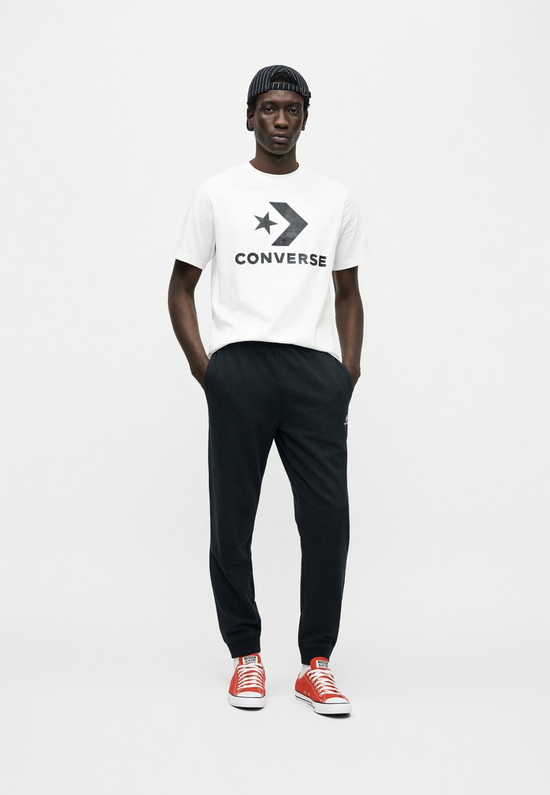 Hvid T-shirt med sort "CONVERSE" logo og stjerne; sorte joggingbukser; røde sneakers med hvide accenter; sort kasket med striber.