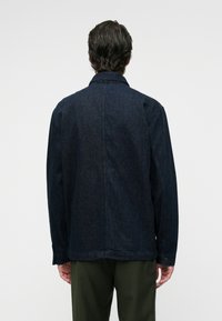 Chaqueta de mezclilla oscura con corte recto, detalles de botones y un cuello clásico. Presenta un diseño limpio en la parte posterior y mangas largas.