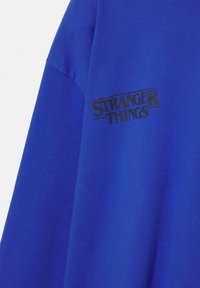 Tricou cu mâneci scurte de culoare albastru regal, din bumbac. Are un logo negru „Stranger Things” pe partea din față. Textură moale și croială casual.