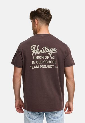 Hombre con cabello corto y castaño que lleva una camiseta marrón oscuro con texto blanco "Heritage Union of '63 & Old School Team Project" en la parte trasera.