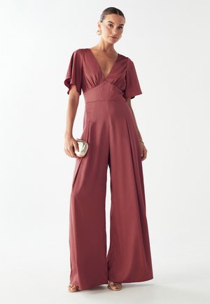 Bordeauxfarbener Jumpsuit mit weitem Bein, kurzen Ärmeln, tiefem V-Ausschnitt, tailliertem Schnitt und Falten. Das Modell hält eine kleine metallische Clutch. Glatte Textur.
