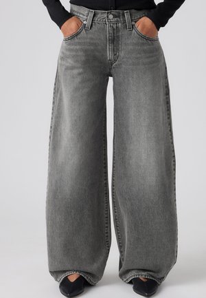 Person, die hoch taillierte, weit geschnittene graue Jeans mit Händen in den Taschen und schwarzen spitzen Schuhen vor einem einfarbigen Hintergrund trägt.