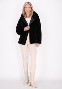 Giacca in finta pelliccia nera con cappuccio, con zip frontale e vestibilità rilassata, indossata sopra una maglietta bianca e abbinata a pantaloni aderenti chiari e stivaletti color crema.