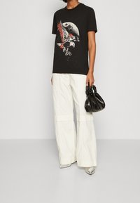 Svart grafisk t-shirt med fågeldesign, ihop med överdimensionerade vita byxor och en liten svart handväska. Texturerat tyg och skarpa skor.