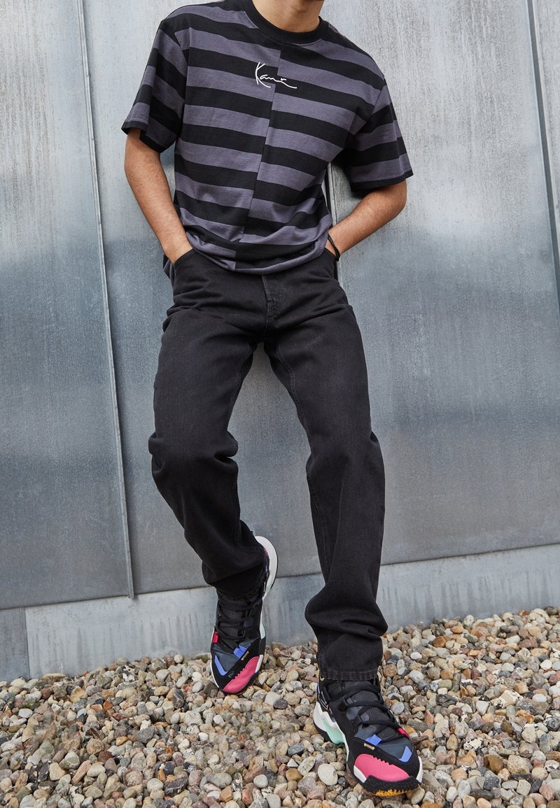 Homme portant un t-shirt rayé noir et gris et un jean noir, debout sur du gravier devant un mur métallique, mains dans les poches.