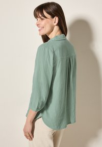 Camicia verde chiaro, con texture, colletto e maniche a tre quarti, caratterizzata da una piega sul retro. Indossata sopra pantaloni beige chiaro.
