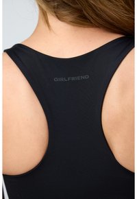 Nærbilde av en kvinnes øvre rygg med svart racerback-topp hvor ordet "GIRLFRIEND" er trykket i grått nær halsen.