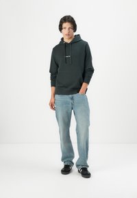 Donkergroene hoodie met een voorzak en zwarte veters, gecombineerd met losse, lichtblauwe jeans en zwarte sneakers. Minimalistisch ontwerp.