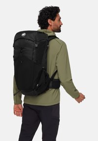 Schwarzer Rucksack mit strukturiertem Design, mehreren Riemen und einer Netzstruktur. Der Benutzer trägt ein olivgrünes Langarmshirt.