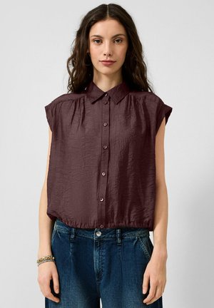 Frau mit langen dunklen Haaren, die eine ärmellose, dunkelviolette, geknöpfte Bluse trägt, in blaue Jeans gesteckt, vor schlichtem Hintergrund.