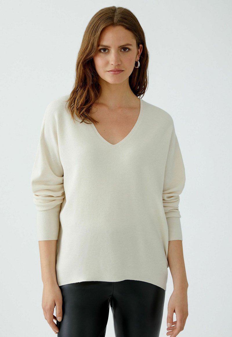 Hellbeige V-Ausschnitt-Pullover mit geripptem Muster und überschnittenen Ärmeln, kombiniert mit schwarzen, figurbetonten Hosen. Aus weichem Strickmaterial, lässige Passform.