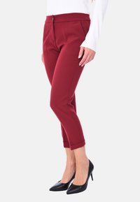 Pantaloni rossi su misura con un design aderente, chiusura frontale con zip e orli arrotolati, abbinati a scarpe nero con tacco alto, che mettono in risalto una silhouette elegante.
