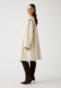 Cappotto in pelliccia bianca con cuciture prominenti, maniche lunghe e vestibilità ampia. Abbinato a stivali in suede marrone alti fino al ginocchio con tacco a spillo.