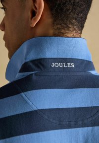 Homme portant un polo rayé bleu et marine avec le col relevé, affichant le logo "JOULES" brodé sur le col arrière.