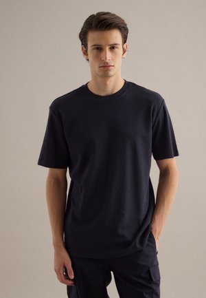 Junger Mann mit dunklem Haar steht frontseitig, trägt ein schlichtes schwarzes T-Shirt mit kurzen Ärmeln und dunkle Hosen vor einem neutralen Hintergrund.