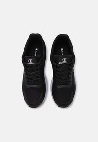 Baskets de sport noires en matière synthétique avec une tige texturée, un col rembourré et des lacets noirs. Elles disposent d'une semelle en caoutchouc blanche et d'un logo.