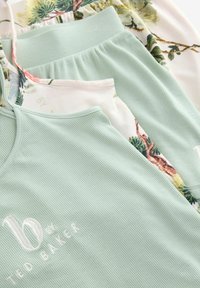 Groene geribbelde tanktop met witte geborduurde tekst, gelaagd over een bloemenprint broek met een elastische tailleband en een zachte textuur.