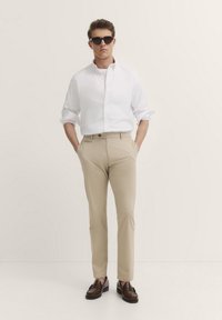 Weißes Hemd mit Knöpfen, beige Chinos, braune Loafers und schwarze Sonnenbrille, vor einem neutralen Hintergrund stehend. Maßgeschneiderte Passform, klassisches Design.