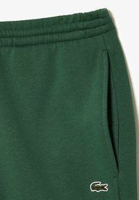 Pantalones de chándal verde oscuro con cintura elástica, bolsillo lateral y pequeño logo de cocodrilo bordado cerca del bolsillo en la pierna derecha.