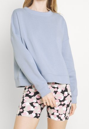 Kvinde iført en lyseblå sweatshirt med lange ærmer og højtaljede cykelshorts med rosa, hvide og sorte abstrakte pletter.