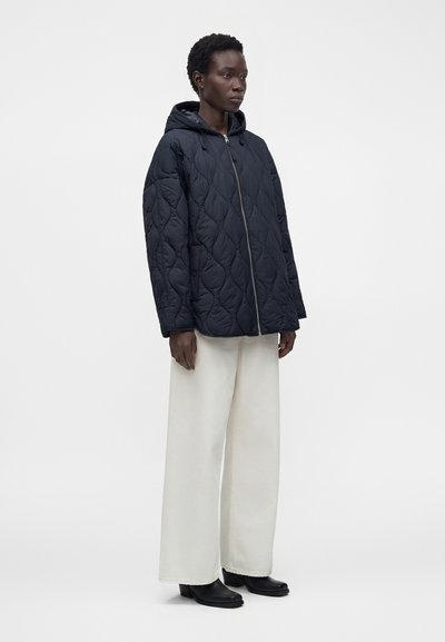 Personne portant une veste noire matelassée à capuche, un pantalon blanc large et des bottines noires, debout devant un fond clair uni.