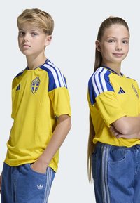 Gula fotbollströjor med blå detaljer, prydda med Sveriges riksvapen, som bärs av två barn. Blå jeans med vita ränder.