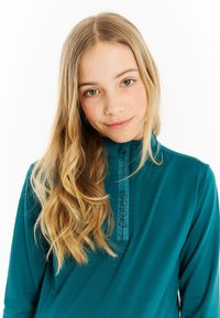 Teal polo s dlhým rukávom s vysokým golierom, vybavený zipsom a čiernym logom na prednej strane. Mäkka tkanina s hladkou textúrou.