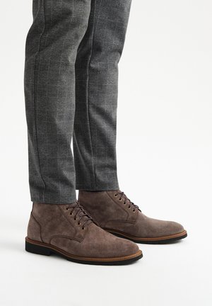 Brązowe zamszowe botki za kostkę z okrągłym noskiem, przeszyciami i metalowymi oczkami na sznurowadła, w połączeniu z szarymi spodniami w wzory.