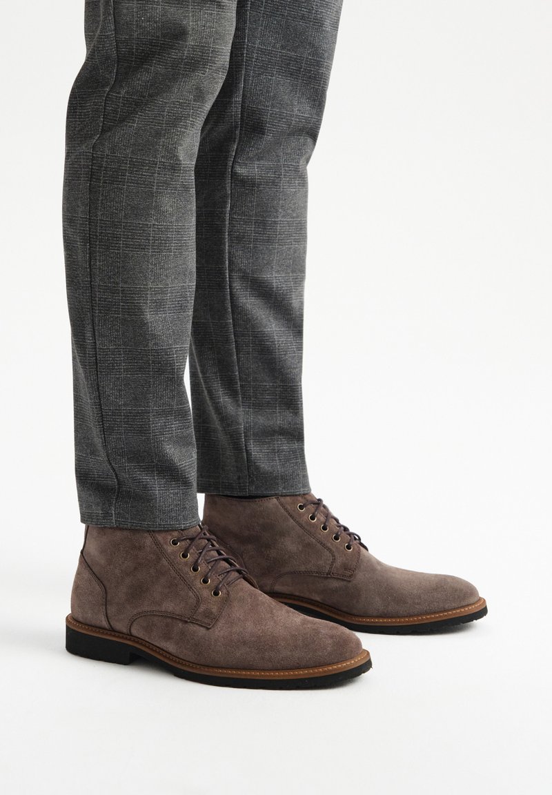 Brązowe zamszowe botki za kostkę z okrągłym noskiem, przeszyciami i metalowymi oczkami na sznurowadła, w połączeniu z szarymi spodniami w wzory.