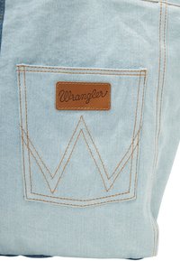 Jasnoniebieska tkanina dżinsowa z tylną kieszenią ozdobioną prostokątną brązową skórzaną naszywką Wrangler oraz kontrastującymi złotymi przeszyciami.