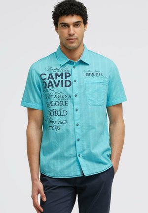 Mann trägt ein kurzärmliges türkisfarbenes Hemd mit schwarzem Aufdruck und marineblaue Hose, steht vor einem einfarbigen Hintergrund.