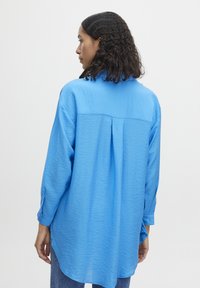 Camicia azzurro chiaro a manica lunga realizzata in tessuto texture. Presenta un yoke posteriore, dettagli con pieghe e polsini con bottoni per una vestibilità regolabile.