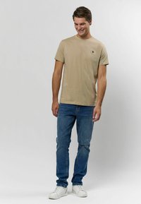 Beige bomullströja med rundad halsringning och liten mörk logotyp. Bärs med blå denimjeans och vita sneakers; avslappnad passform.