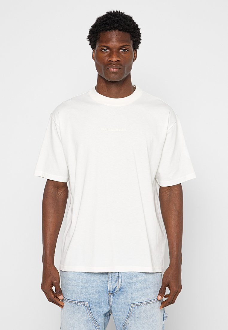 Man met kort krullend haar, gekleed in een eenvoudig wit oversized T-shirt en lichtblauwe spijkerbroek, staand met het gezicht naar voren.