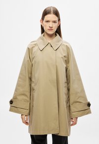 Bézs színű, túlméretezett kabát, nagy gallérral, széles harang alakú ujjakkal és gombos mandzsettával. Oldalzsebekkel és sima tapintású anyaggal rendelkezik.