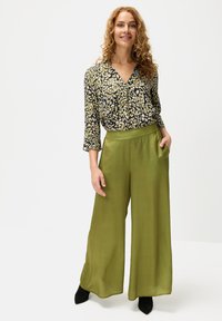 Die Bluse hat ein florales Muster in Gelb und Grün auf einem dunklen Hintergrund; grüne weite Hosen mit seitlichen Taschen und glatter Textur.