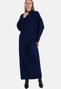 Abito in maglia blu navy con colletto alto, maniche lunghe e dettaglio in vita annodato; lungo fino al pavimento, con una texture liscia e un design minimalista.