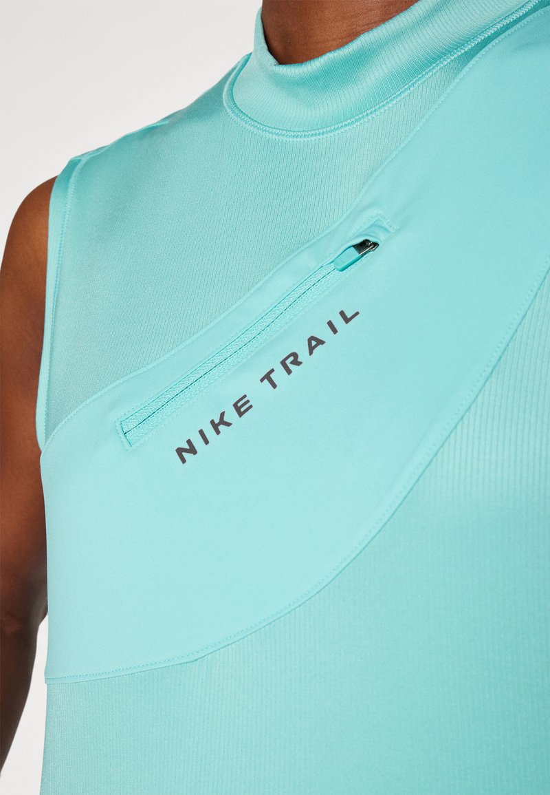 Un haut de sport sans manches, de couleur turquoise clair, avec un col montant, présentant une poche zippée et le texte "NIKE TRAIL" imprimé en noir.
