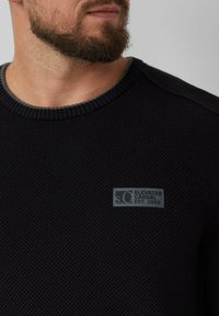 Homme barbu portant un pull en texture noir avec un patch gris portant l'inscription "s.Oliver Elevated Casual Est. 1969" sur la poitrine.