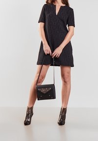 Vestito nero a maniche corte con scollatura a V, in tessuto testurizzato, impreziosito da una borsa nera. Il modello indossa stivali metallici alla caviglia con un motivo distintivo.