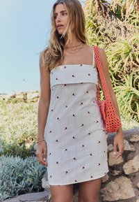 Weißes, eng anliegendes Kleid mit dünnen Trägern, verziert mit roter Blumenstickerei und vertikalen Streifen. Akzentuiert mit einer strukturierten rosa Handtasche.