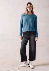 Hellblauer strukturierter Pullover, dunkle locker geschnittene Jeans, weiße Sneakers mit grauen Socken. Einfaches Design und lässige Passform. Neutraler Hintergrund.
