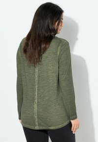 Vrouw met lang donker haar, gekleed in een olijfgroen overhemd met lange mouwen en zwarte broek, staat met de rug naar de kijker gericht tegen een eenvoudige witte muur.